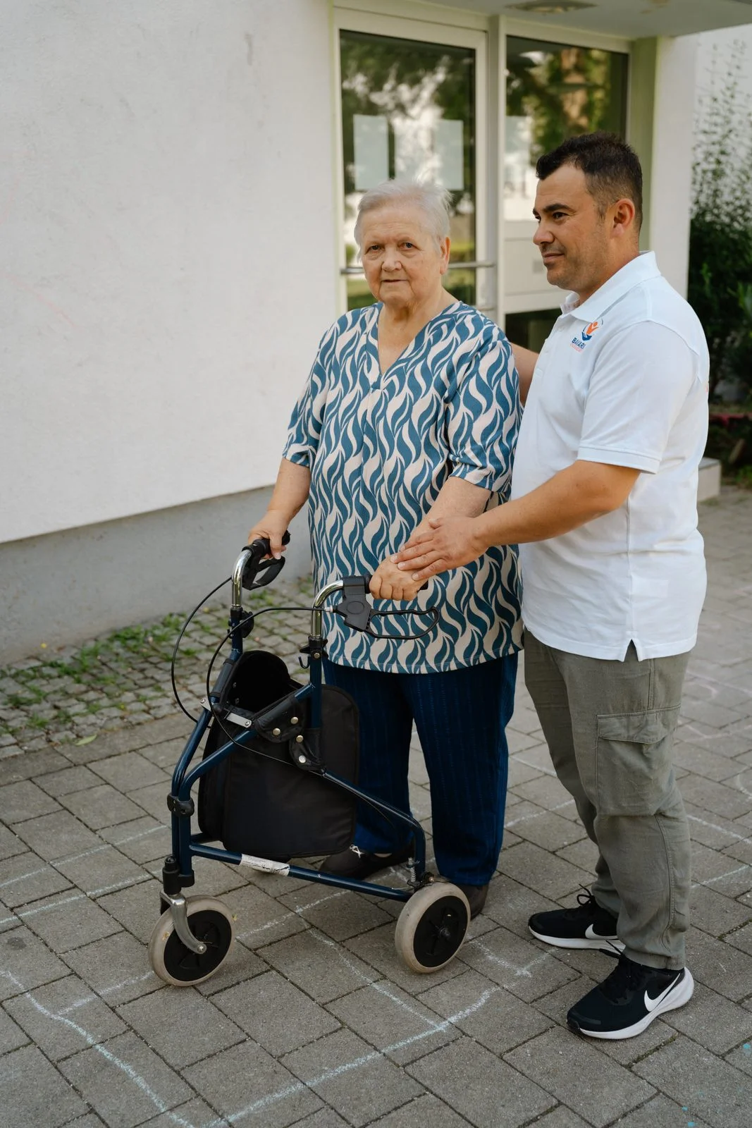 Pflegekraft von Bavaria Vitalpflegedienst begleitet eine Seniorin mit Rollator an der Haustür